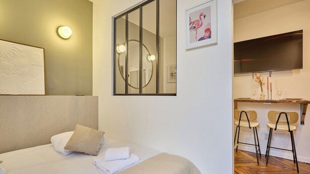 A hotel room with a bed, a desk with two chairs, a television, and a wall-mounted mirror. - Charming Apartment - Paris Pere Lachaise (Paris)