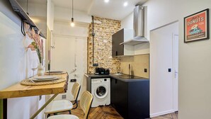A compact kitchen with a brick wall, a washing machine, and a dining area with a wooden table and chairs. - Charming Apartment - Paris Pere Lachaise (Paris)