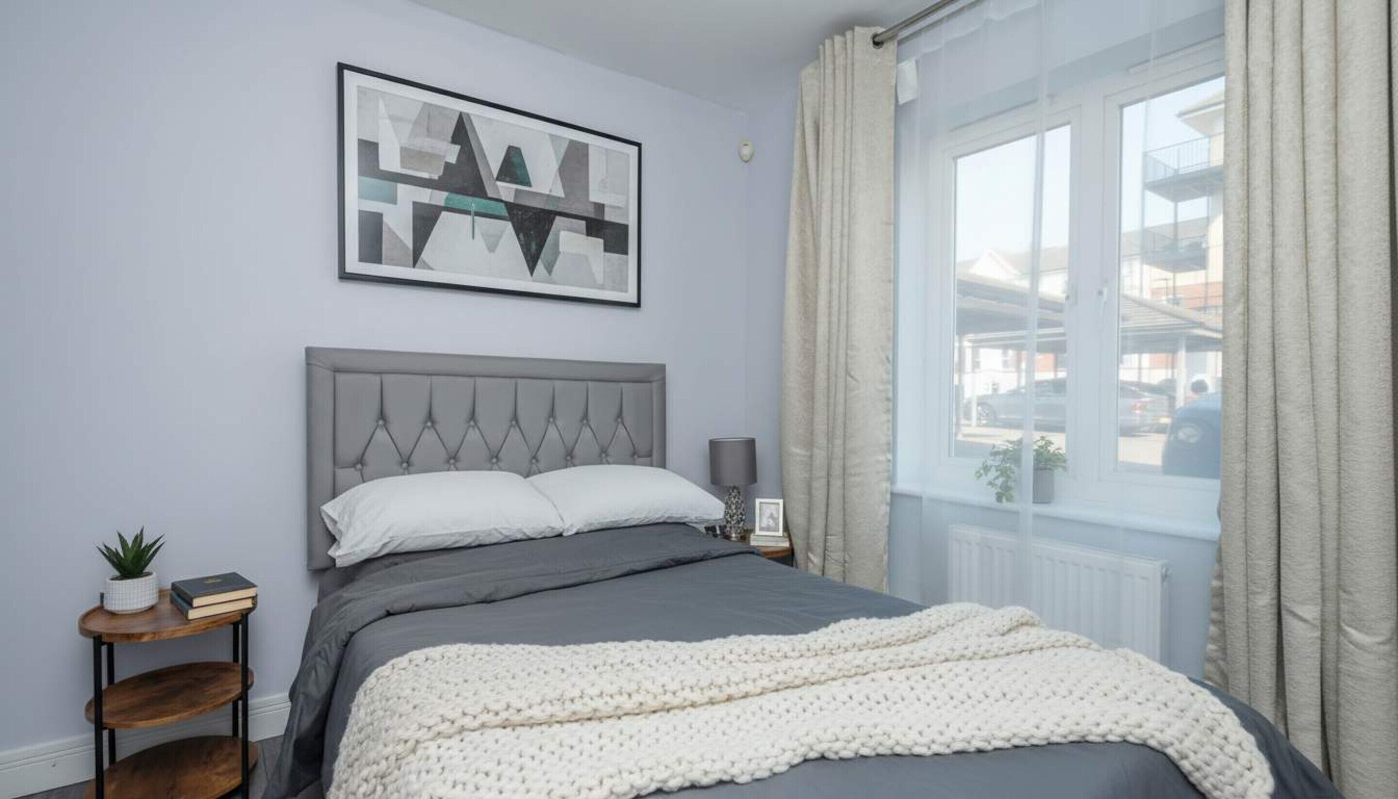 Een slaapkamer met een grijs gestoffeerd bed, een kunstwerk met abstracte vormgeving erboven, een nachtkastje met een lamp en een raam met gordijnen.