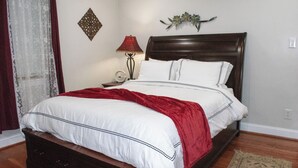 A neatly made bed with white bedding and a red throw blanket, a bedside lamp, a decorative wall piece, and a small rug on a wooden floor.