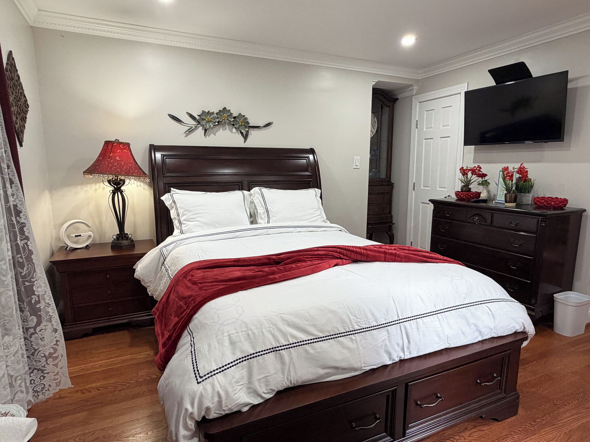 A bedroom with a wooden bed, white bedding, a red throw blanket, a nightstand with a lamp, a dresser, and a TV on the wall.