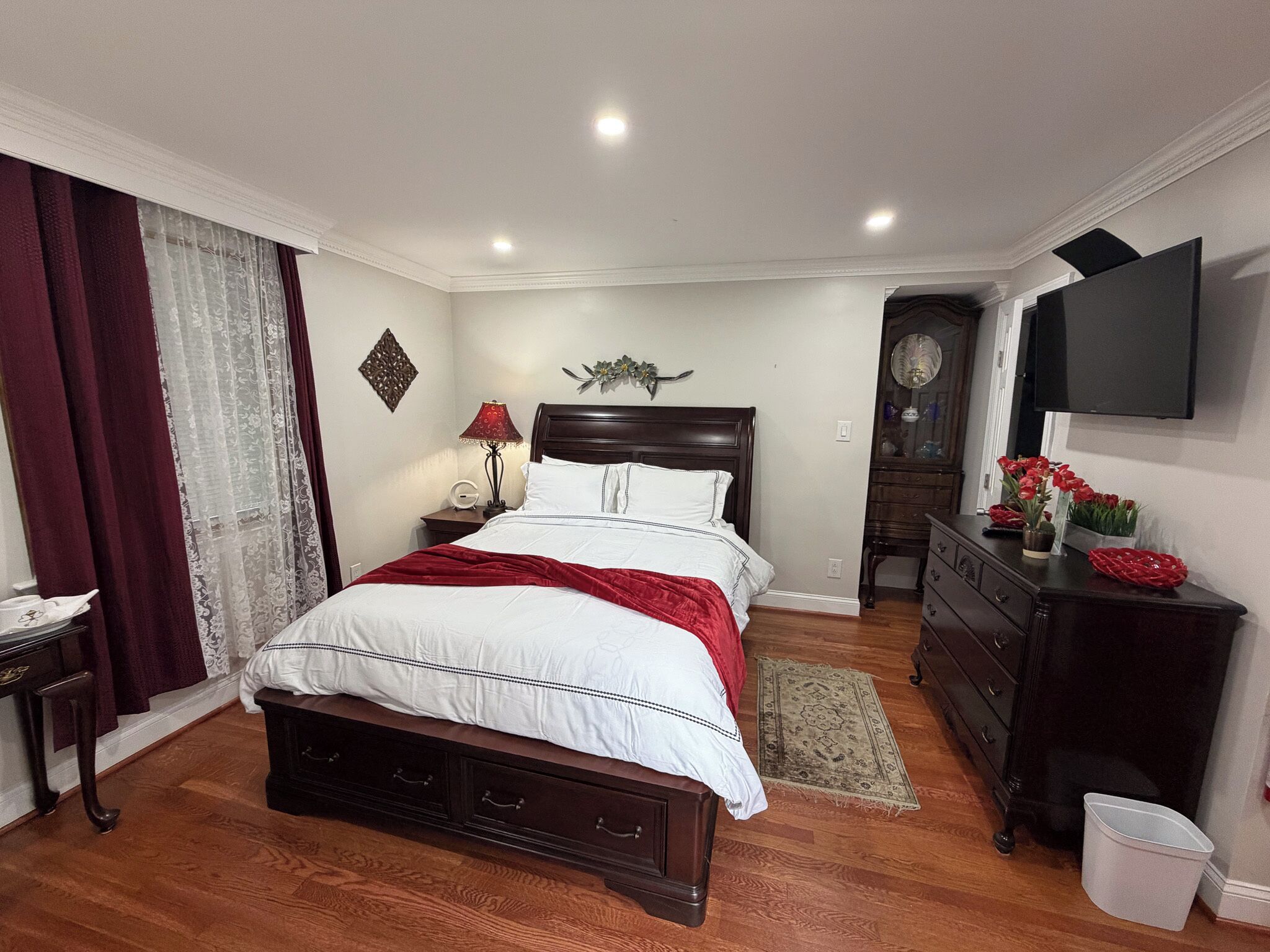 A bedroom with a bed, dresser, nightstand, lamp, and a TV on the wall.