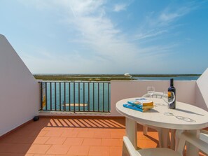 A balcony with a table, a bottle, and glasses, overlooking a body of water and distant land. - Apartamento T0 Vista Mar Vista Ria Formosa (Cabanas de Tavira)