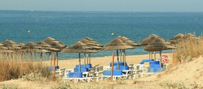Een strand met rieten parasols en blauwe ligstoelen.