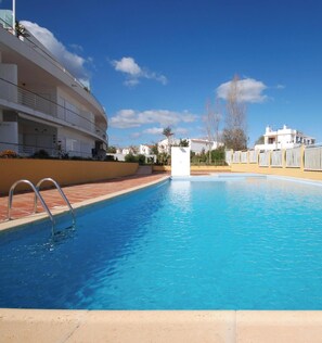 Una piscina con agua cristalina azul, rodeada por un edificio con balcones y un cielo despejado.