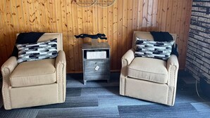 Zwei Sessel mit gemusterten Kissen, ein kleiner Holztisch mit einem dekorativen Gegenstand und eine Holzwand mit Backsteinmuster auf der rechten Seite.