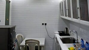 Una cocina compacta con paredes de azulejos blancos, estufa integrada, un lavamanos y una zona de comedor con sillas de plástico.