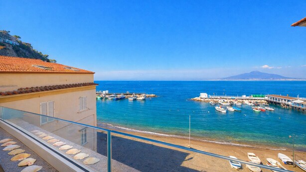 Vista sulla costa con un balcone affacciato sul mare, barche ormeggiate al molo e una montagna sullo sfondo.