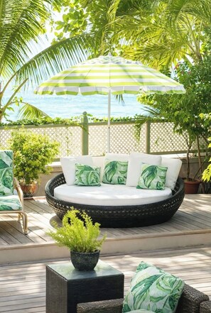 A round outdoor seating area with a white cushion and green leaf-patterned pillows, surrounded by a lattice fence and palm trees. - Cashmere Properties, Barbados Beachfront Villa, 3BR+Cottage-4BR option (Bridgetown)