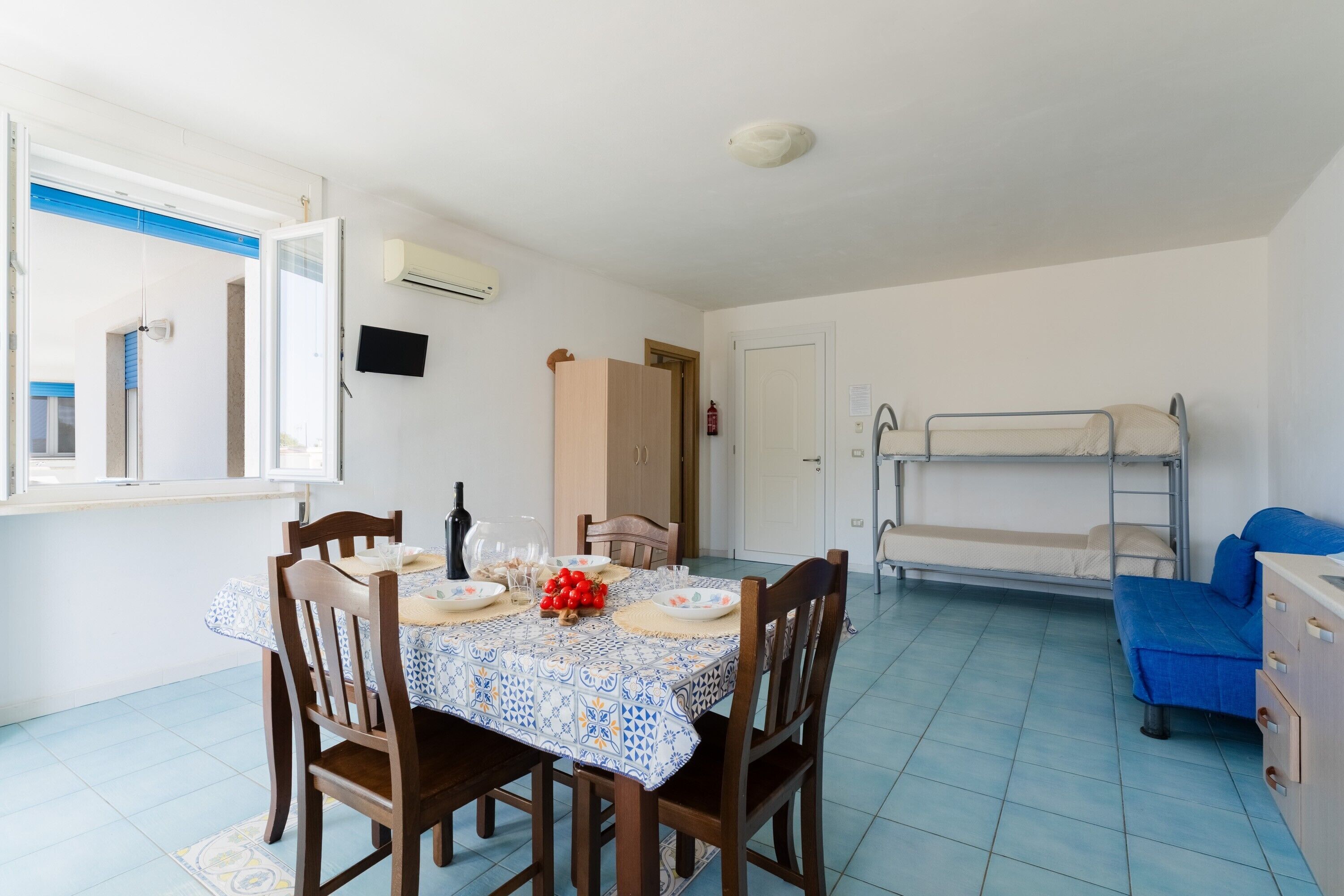 A room with a dining table set for four, a bunk bed, a TV mounted on the wall, and a door leading to another room.