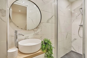 Une salle de bain avec un miroir rond, un lavabo blanc, une douche et une plante en pot.