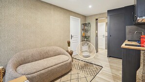 Ein modernes Wohnzimmer mit einer Couch, einem Sessel, einem kleinen Tisch und einer Küchenzeile mit Schränken und einer Arbeitsplatte.