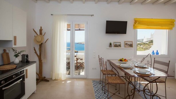Una cocina con estufa, una mesa de comedor para cuatro personas, un televisor montado en la pared y vista al mar a través de una puerta.