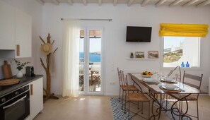 Une cuisine équipée d’une cuisinière, une table à manger pour quatre personnes, un téléviseur fixé au mur et une vue sur la mer par une porte.