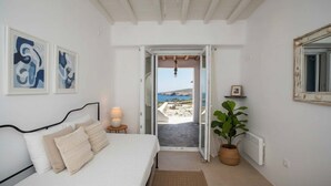 Un dormitorio con cama, lámpara de noche, planta en maceta y vista al mar a través de una puerta abierta.