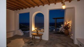 Una habitación con piso de piedra, techo de madera, una pequeña mesa con un florero, y vista al mar a través de una ventana en arco.