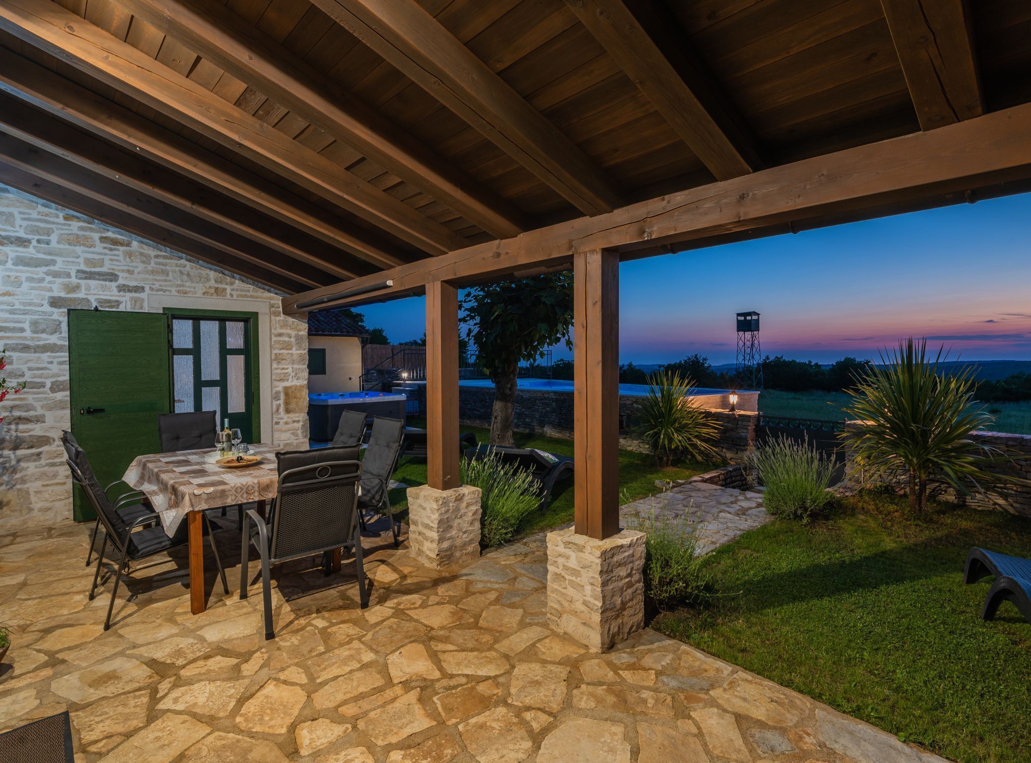 A stone patio with outdoor seating, a hot tub, and a view of the landscape at dusk.