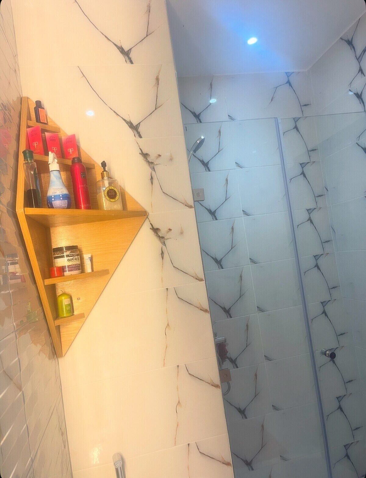 A bathroom with a wooden shelf holding toiletries, a cracked marble wall, and a shower area.