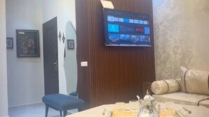 A modern living room with a flat-screen TV mounted on a wooden panel, a dining table with a tray of tea, and a sofa with patterned cushions. - Modern Wi-Fi, Agadir, 10 min Beach, Netflix, IPTV, First floor. 93m2. Balcony. (Agadir)