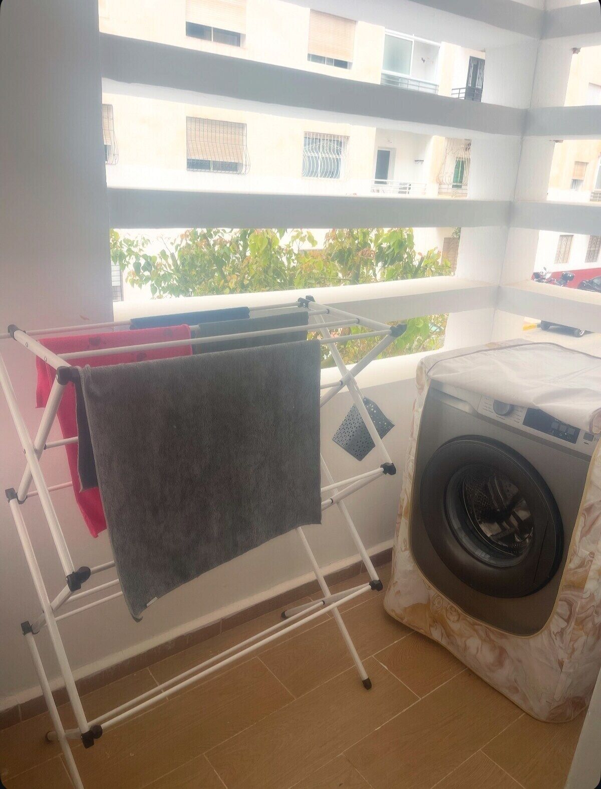 Een balkon met een droogrek, een wasmachine en uitzicht op gebouwen en groen.