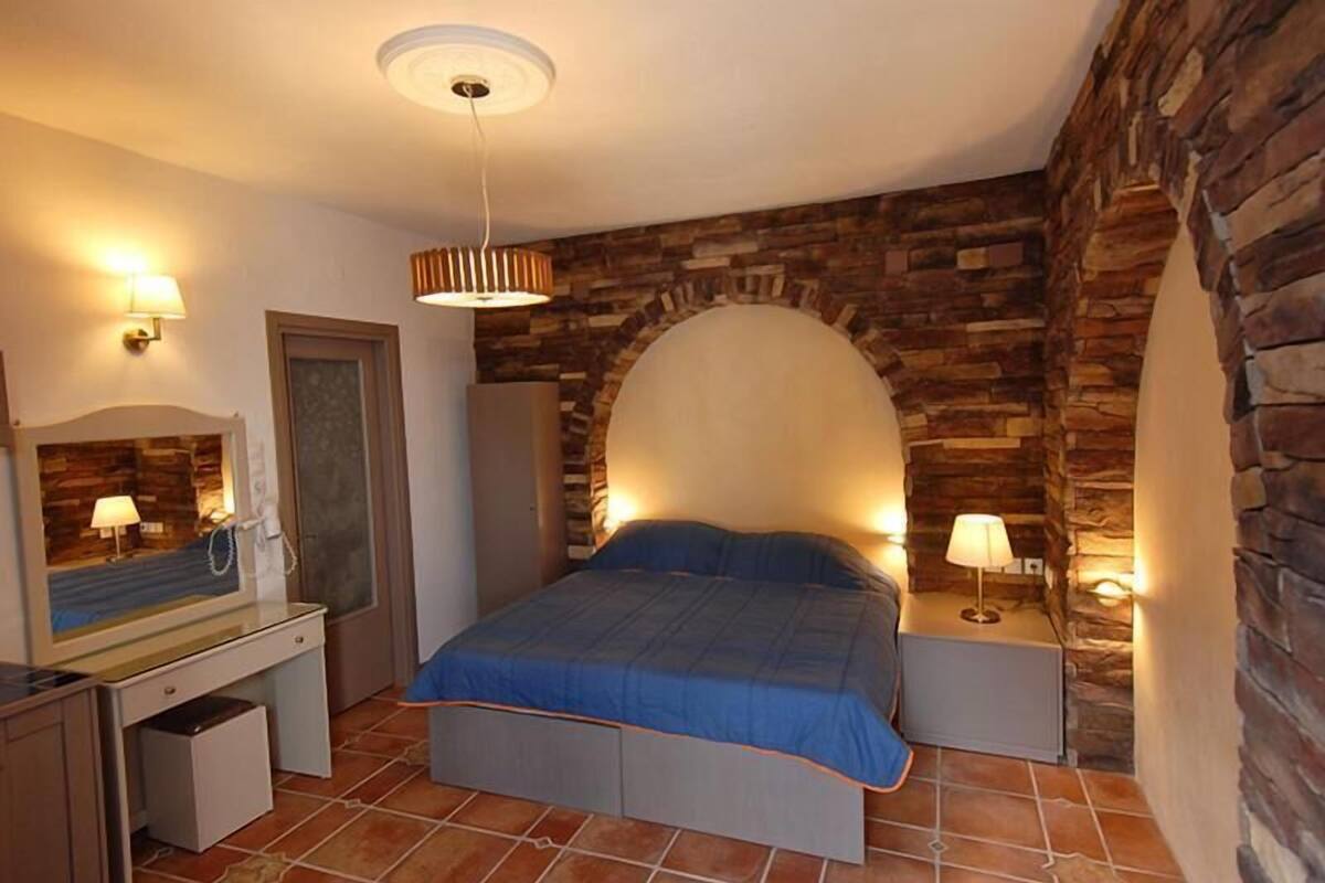 A bedroom with a stone wall arch, a bed with a blue bedspread, a desk with a mirror, and two bedside lamps.