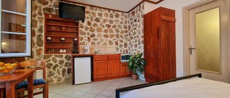 A bedroom with a bed, a small kitchenette, a dining area with a table and chairs, and a stone wall.