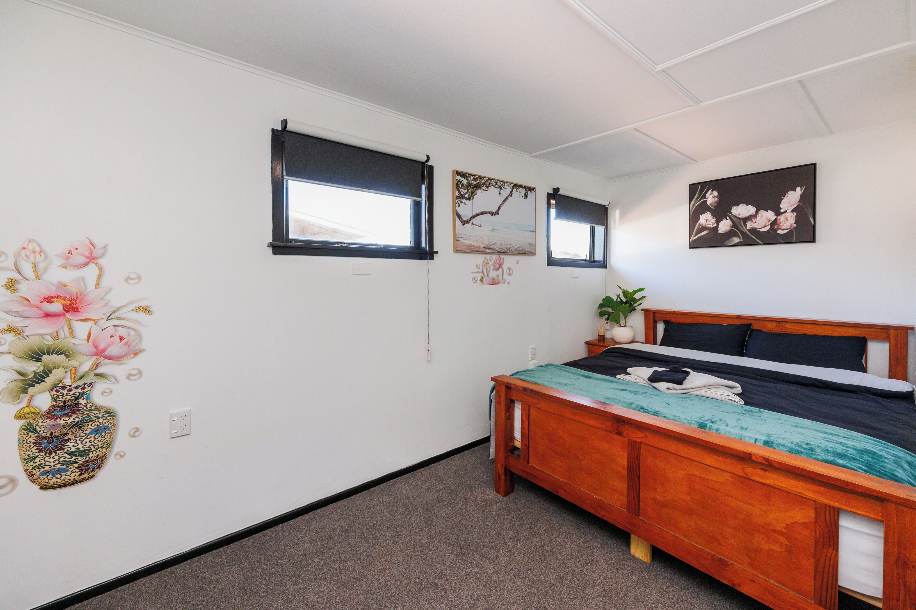 A bedroom with a wooden bed, a window, a wall-mounted picture, and a vase with flowers.