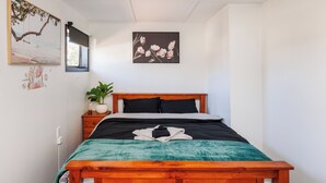 Ein Schlafzimmer mit einem hölzernen Bett, einer türkisfarbenen Decke, einem schwarz-weißen Kissenpaar und einem gerahmten Blumenbild, sowie einer Zimmerpflanze.