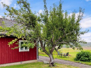 A red wooden building with a window, a tree in front, and a table with chairs in the background.