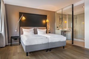 A modern hotel room with a large bed, bedside lamps, a glass shower enclosure, and a view into the bathroom. - Garner Hotel Friedrichshafen By IHG (Friedrichshafen)