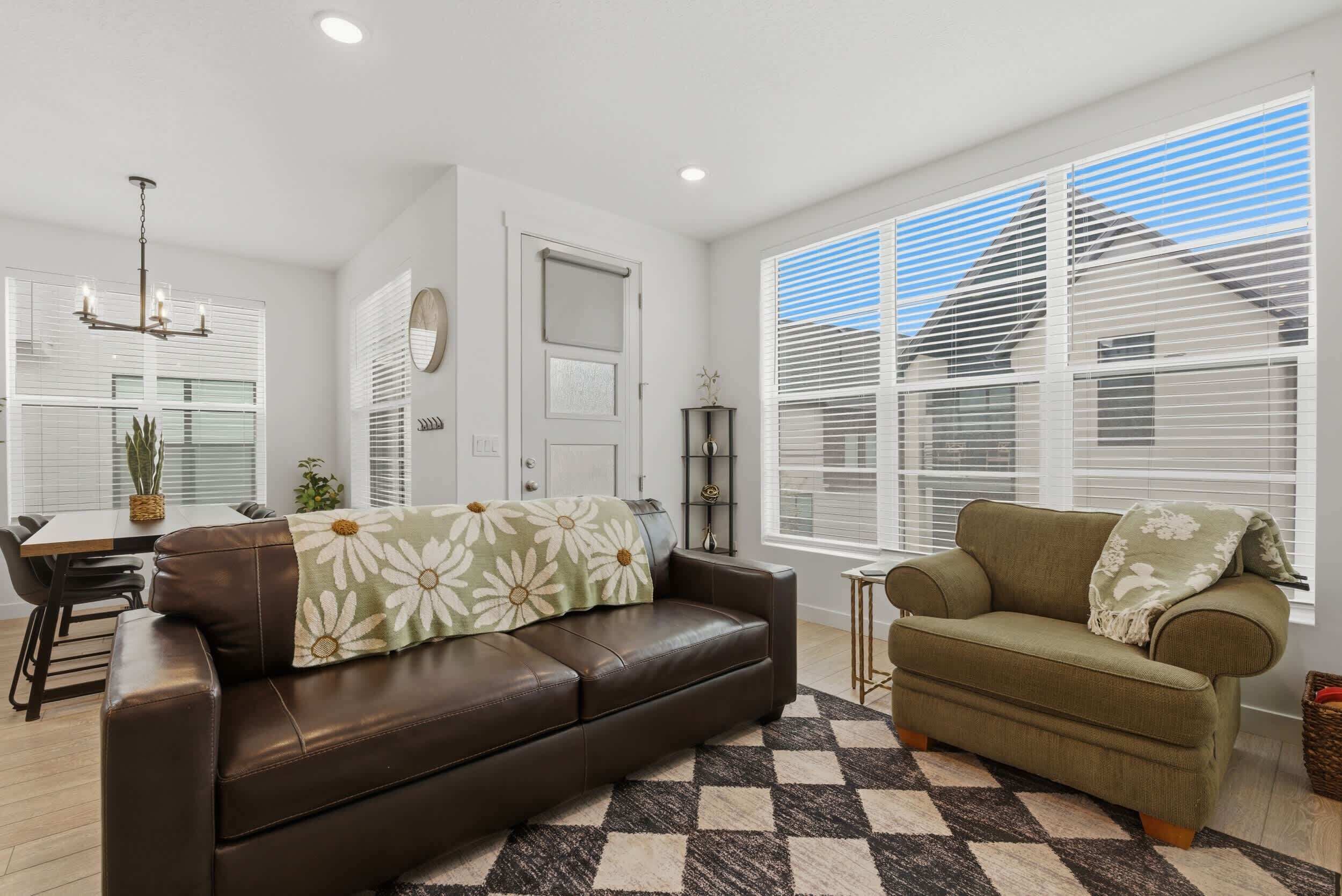 A modern living room with a brown leather sofa, a green armchair, a dining area with a table and chairs, and large windows with blinds.