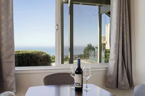 Een tafel met een fles wijn en twee glaasjes, een raam met gordijnen en uitzicht op zee.
