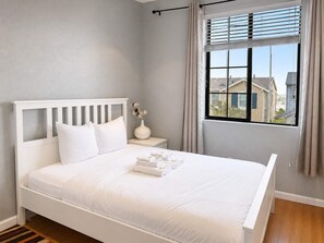 A bedroom with a white bed, a nightstand, a vase with flowers, and a window with curtains.