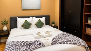 Un letto ben rifatto con una coperta a fantasia, due cuscini decorativi, un set di asciugamani piegati, un quadro astratto appeso al muro, una pianta in vaso e un armadio in legno scuro.