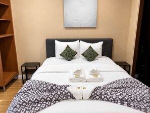 Een net opgemaakt bed met een gebreide deken, twee decoratieve kussens en opgevouwen handdoeken op het hoofdbord.
