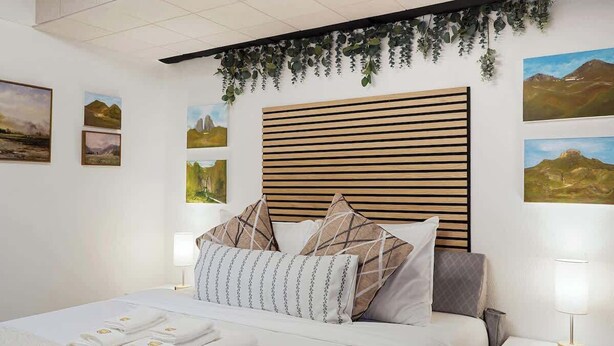 Hotellihuone, jossa on sänky, yöpöytälamput, sängynpääty ja seinätaideteoksia.