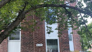 A brick building with a white door, a visible address sign, and a small garden area with potted plants and a tree. - Cozy 1-bedroom studio in pleasant Crofton with AC (Crofton)