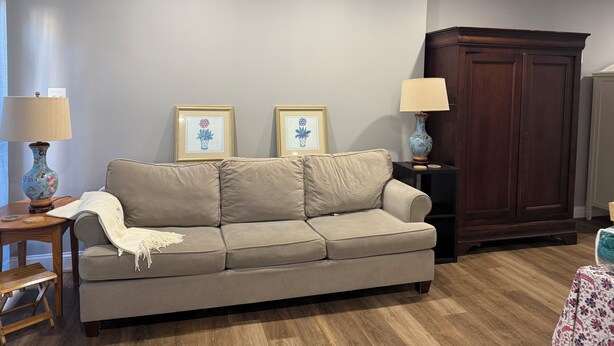 A living room with a beige sofa, two framed floral pictures, a wooden cabinet, and a small table with a lamp. - Cozy 1-bedroom studio in pleasant Crofton with AC (Crofton)