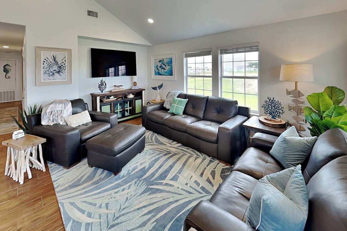 A modern living room with a large sectional sofa, a coffee table, and a TV. There are two windows with blinds, a fireplace, and a large rug.