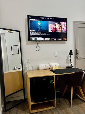 A room with a TV mounted on the wall displaying various shows, a desk with a chair, a small table with a bottle and glasses, and a black device on the floor. - Jazz & Stay (Guadalajara)