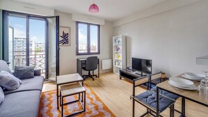 En modern lägenhet med en soffa, ett matbord, en TV, en bokhylla och en balkong med utsikt över staden.
