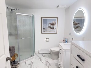 Un baño con mampara de vidrio, inodoro, lavamanos con espejo y un cuadro en la pared.