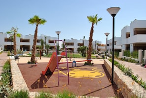 Un parque infantil con tobogán, columpios y estructura de escalada, rodeado de palmeras y edificios modernos.