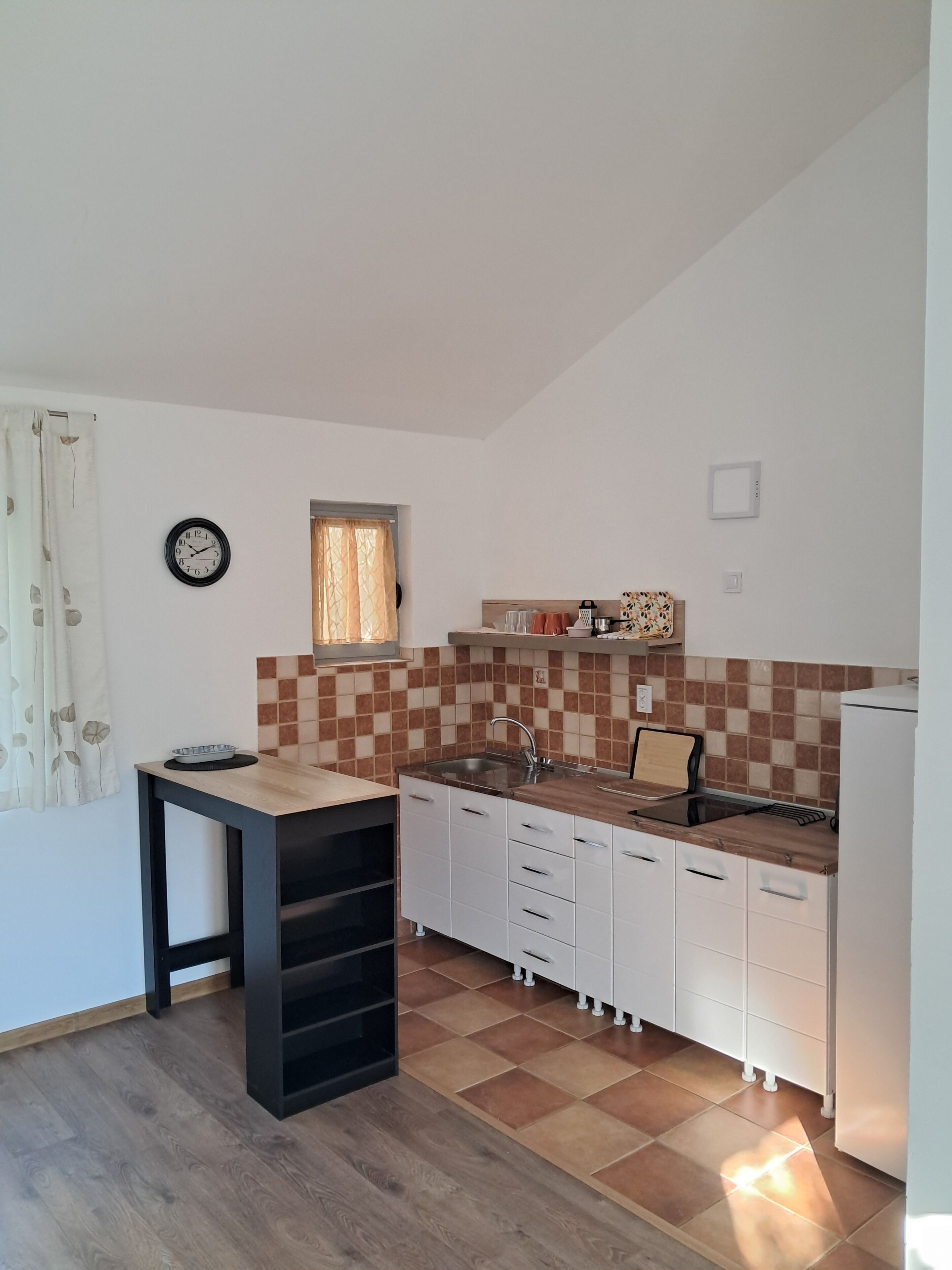 A compact kitchen with a sink, countertop, and cabinets. There is a clock on the wall and a window with curtains.