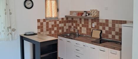 A compact kitchen with a sink, countertop, and cabinets. There is a clock on the wall and a window with curtains.