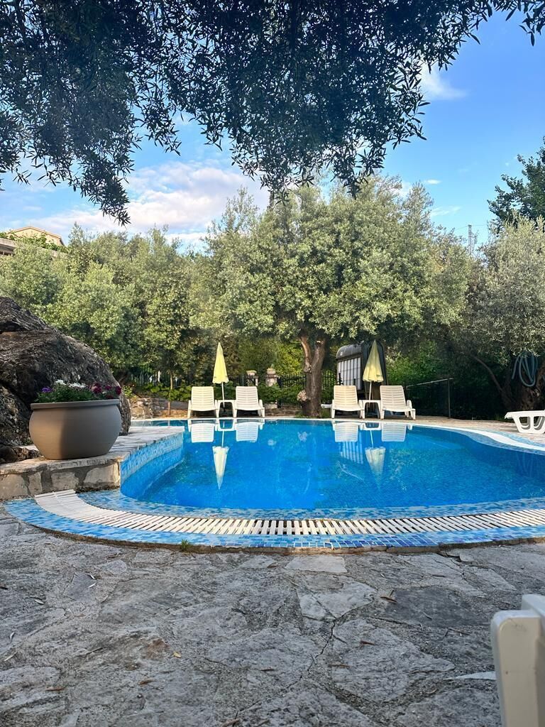Una piscina con lettini e ombrelloni, circondata da alberi e un patio in pietra.