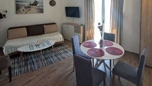 Ein Wohnzimmer mit einer Couch, einem Esstisch mit Stühlen, einem Fernseher und einem Fenster mit Vorhängen.