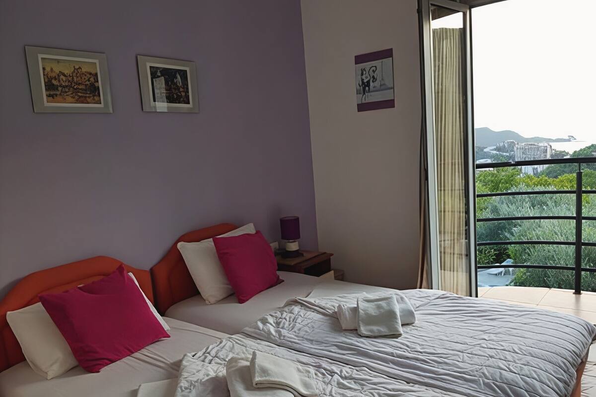 A bedroom with two beds, a nightstand, a lamp, and a window with a view of greenery and buildings.