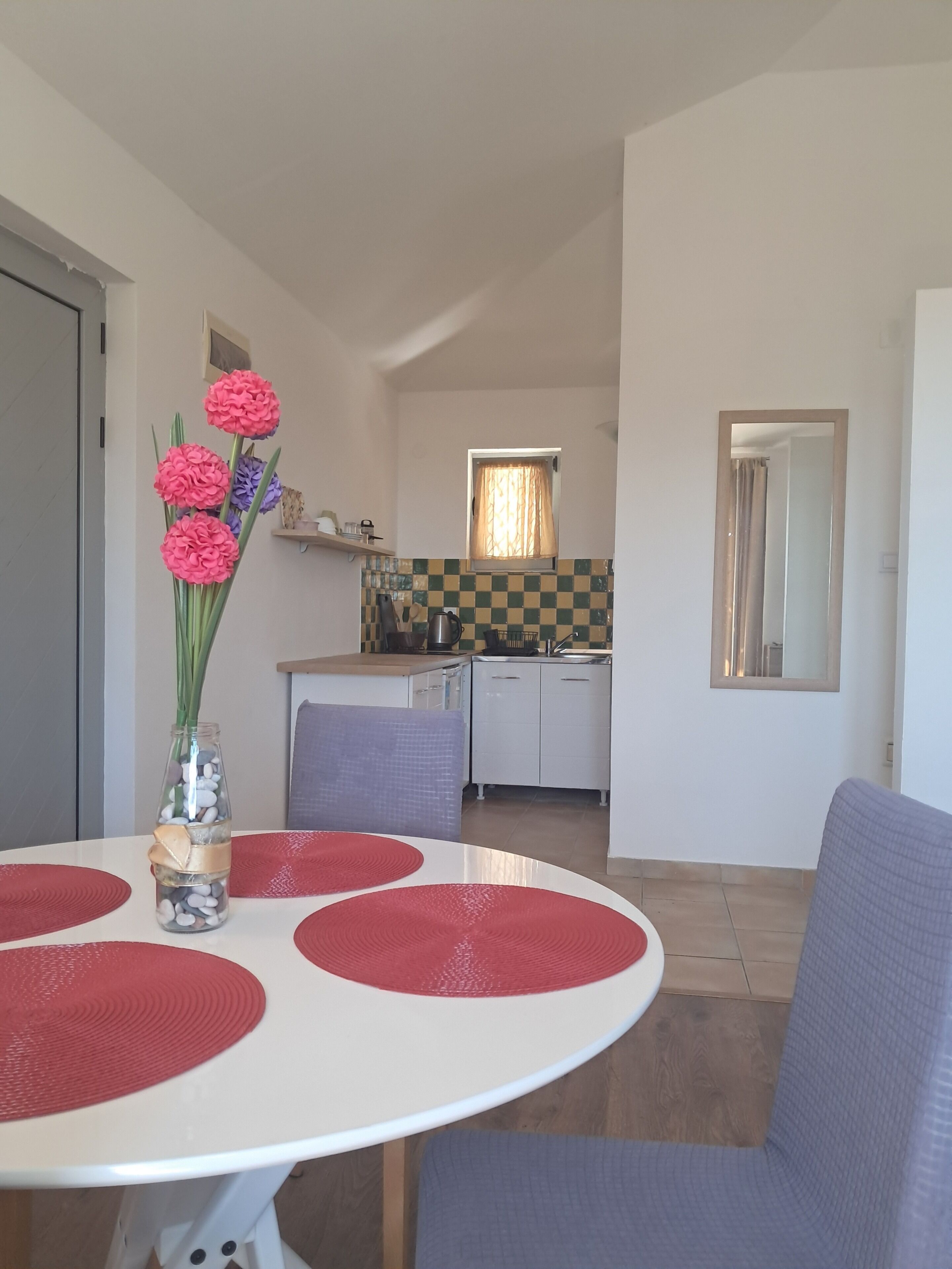 A dining area with a white table, red placemats, and a vase with flowers. A kitchen is visible in the background with a window and a mirror.