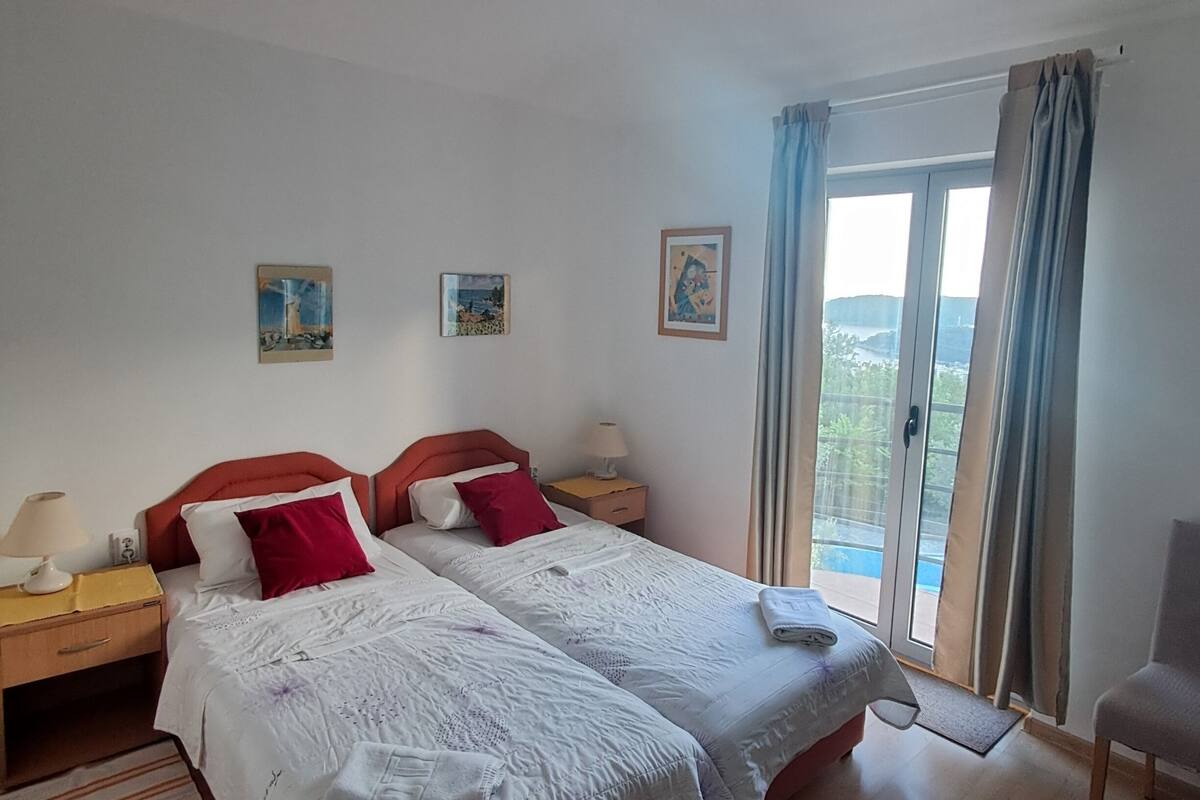 A double bed with white bedding and red pillows, a nightstand with a lamp, and a view of the outdoors through a glass door.